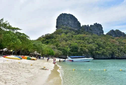 Colorful kayaks on the beach with speedboats anchored in the clear blue water at Angthong National Marine Park.