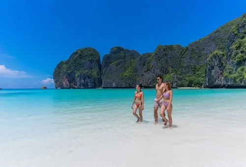 Group of travelers walking along the shoreline at Maya Bay with crystal-clear water and towering cliffs in the background.